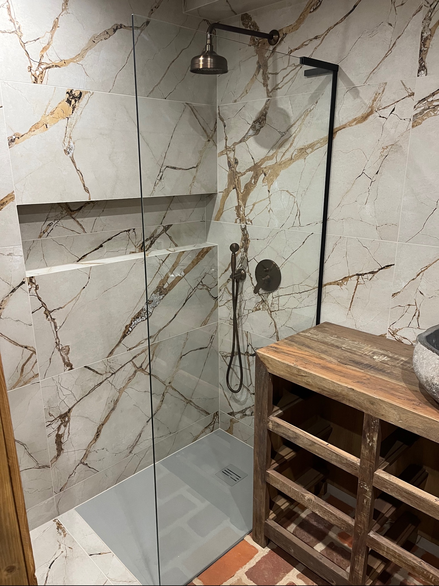 Kabina prysznicowa walk-in z marmurowymi płytkami w odcieniach bieli i złota, wykonana w Gniewinie. Widoczna deszczownica w kolorze starego złota, szklana ścianka oraz drewniana szafka pod umywalkę.