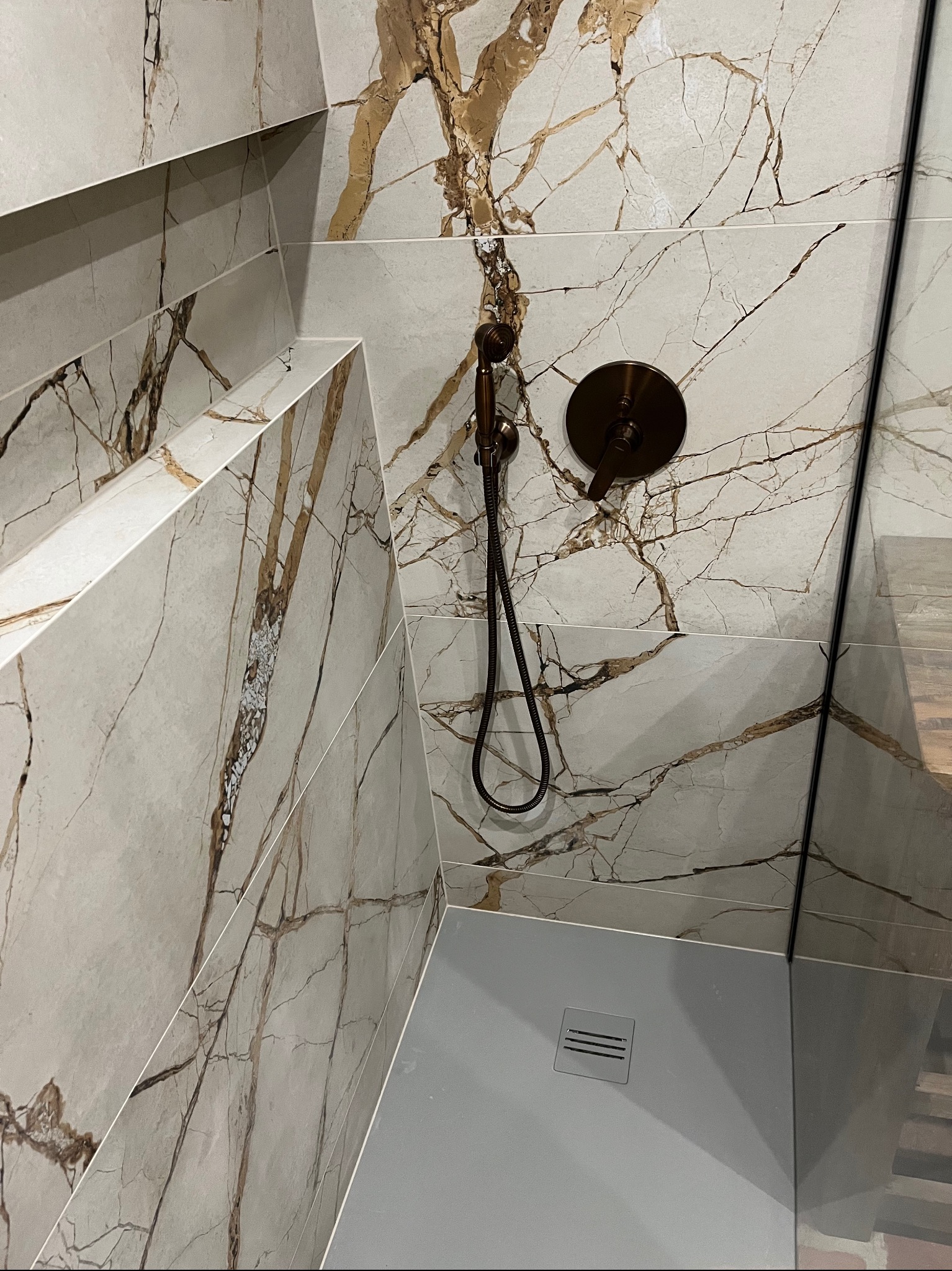 Kabina prysznicowa wykończona płytkami imitującymi marmur z brązowymi żyłami, wyposażona w deszczownicę w kolorze starego złota i odpływ liniowy.