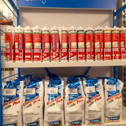 Półka sklepowa z materiałami budowlanymi firmy Mapei: tuby z silikonem Mapesil AC i worki z fugą cementową Ultracolor Plus, ułożone rzędami.