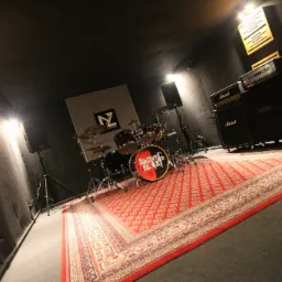 Wnętrze pomieszczenia wygłuszonego panelami akustycznymi, z zestawem perkusyjnym z logo 'Synthetic Blast' na bębnie basowym, wzmacniaczami Marshall i Mesa oraz czerwonym dywanem orientalnym.