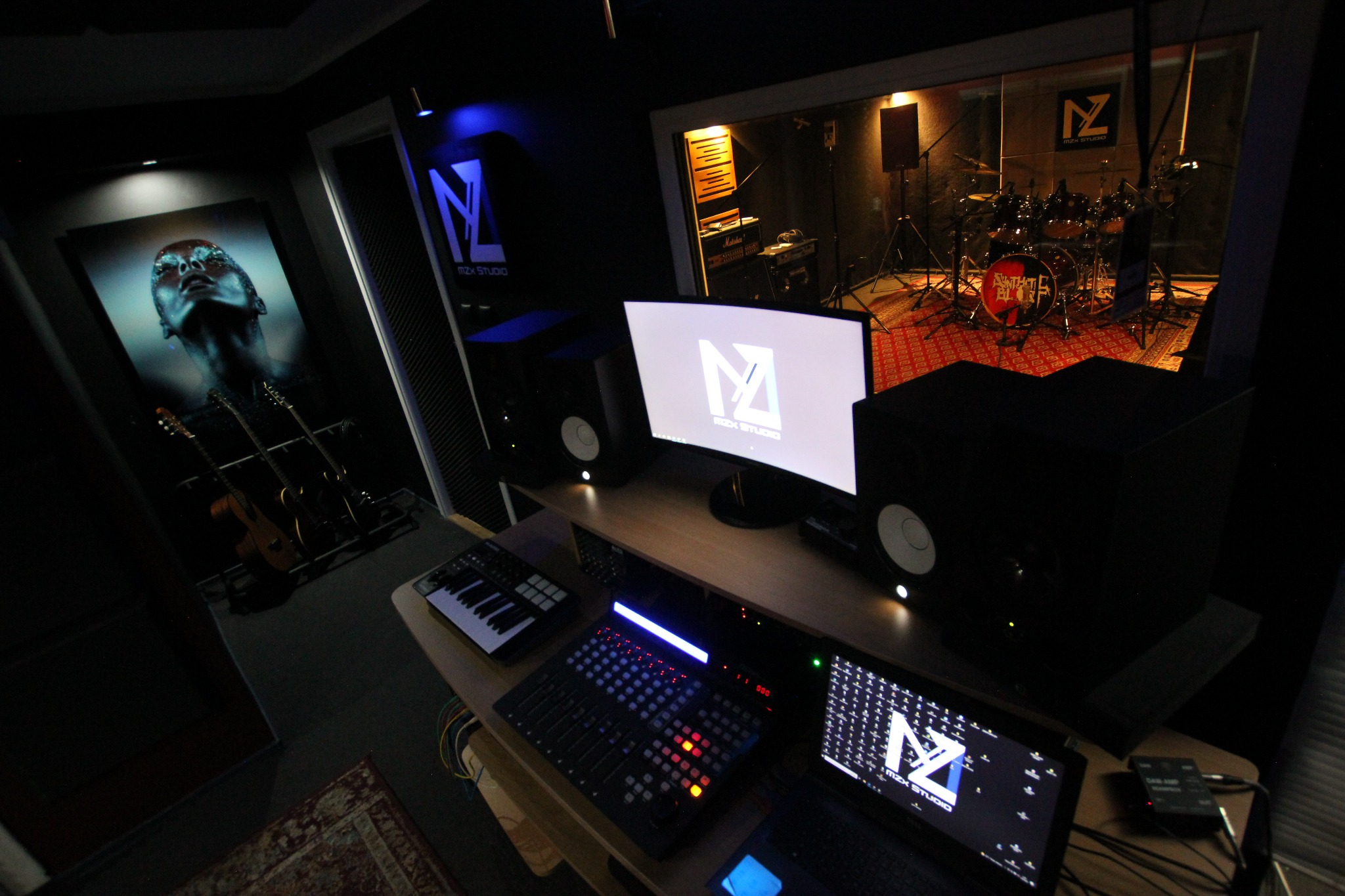 Profesjonalne pomieszczenie do nagrywania z widocznym zestawem perkusyjnym za szybą, mikserem, monitorami studyjnymi, klawiaturą MIDI i gitarami w tle.