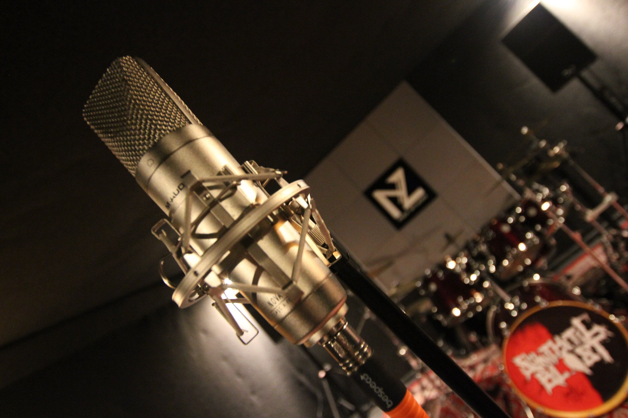 Pojedynczy mikrofon pojemnościowy M-Audio Nova w uchwycie antywibracyjnym, ustawiony pod kątem, z widocznym logiem zespołu na bębnie w tle studia nagrań.