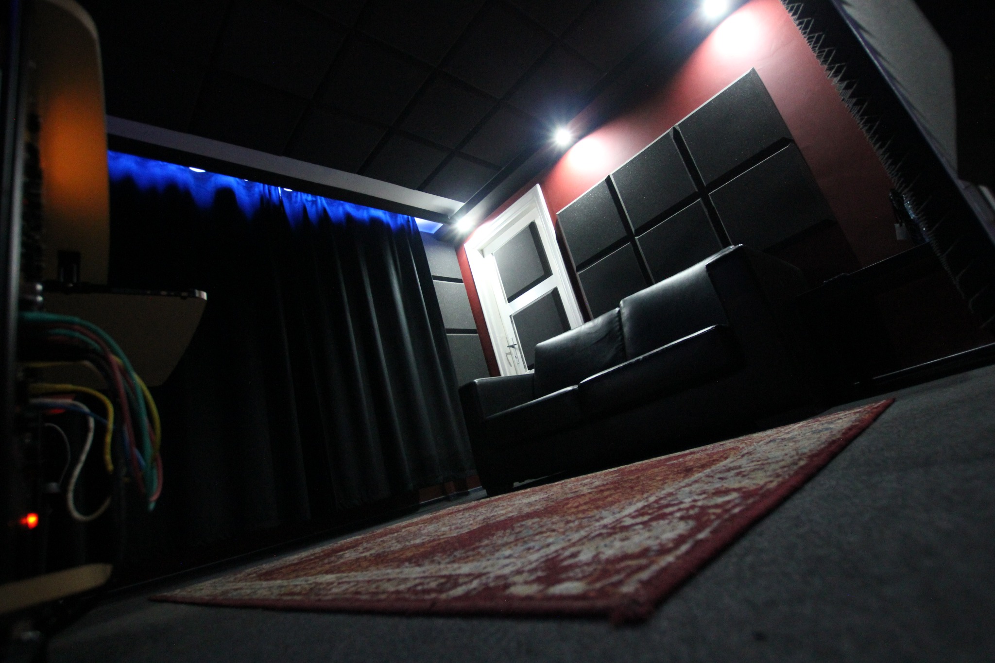 Wnętrze wyciszonego pokoju z czarną sofą, ciemną kotarą z niebieskim podświetleniem, panelami akustycznymi na ścianie i wzorzystym dywanem, widziane z niskiej perspektywy.