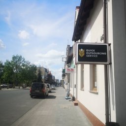 QUICK OUTSOURCING SERVICE SPÓŁKA Z OGRANICZONĄ ODPOWIEDZIALNOŚCIĄ - Zewnętrzny szyld firmy 'Quick Outsourcing Service' zamontowany na elewacji budynku przy chodniku i ulicy w słoneczny dzień.