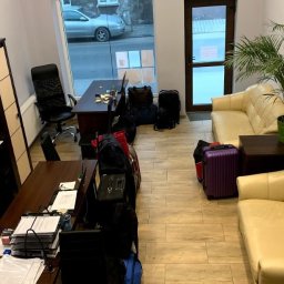 QUICK OUTSOURCING SERVICE SPÓŁKA Z OGRANICZONĄ ODPOWIEDZIALNOŚCIĄ - Biuro z dwoma biurkami, stosem dokumentów, krzesłem biurowym, regałem, sofą, rośliną doniczkową i bagażami, sugerujące tymczasowe miejsce pracy.