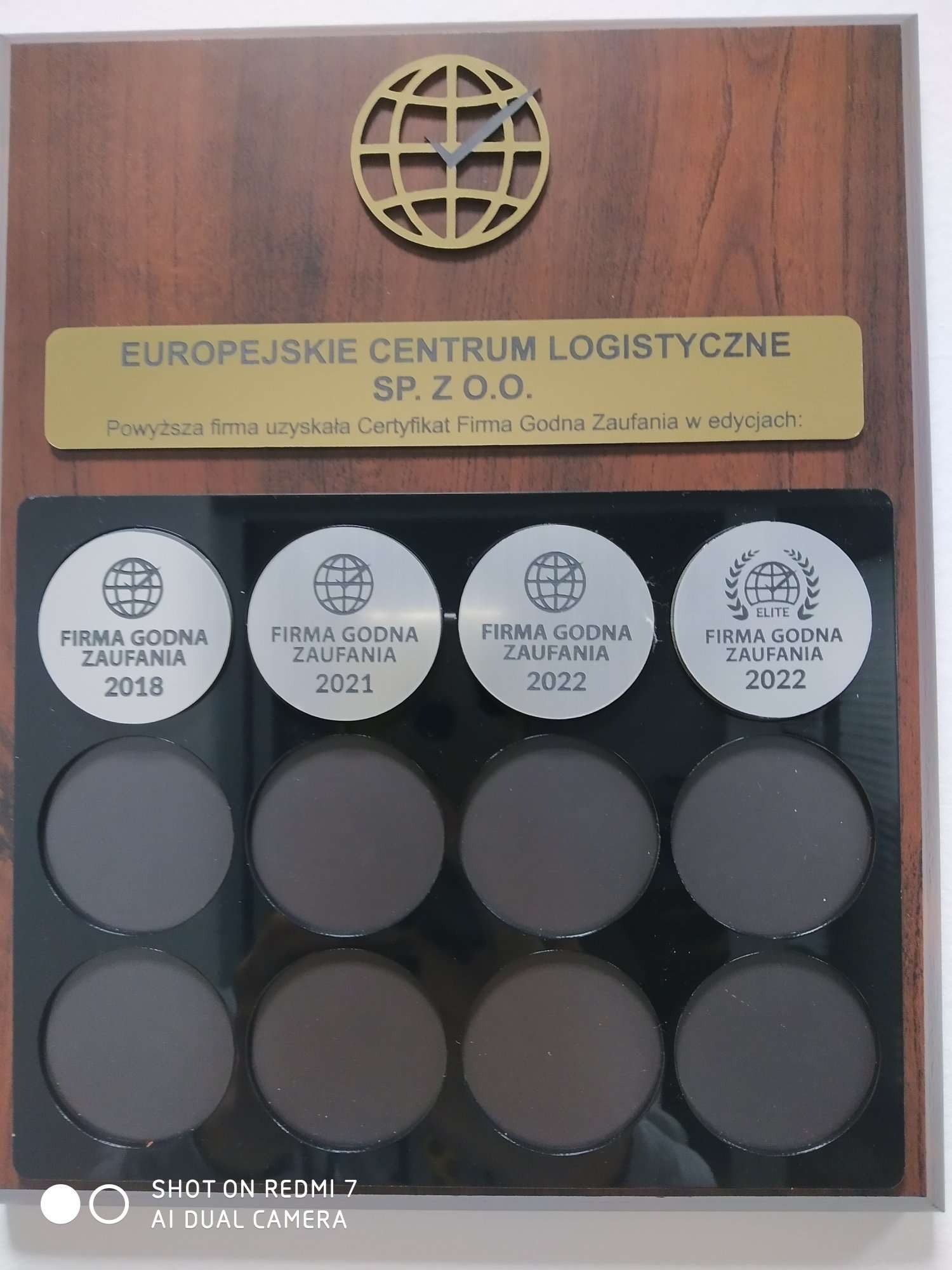Tablica z certyfikatami 'Firma Godna Zaufania' dla Europejskiego Centrum Logistycznego Sp. z o.o. z lat 2018, 2021 i 2022, w tym wersja Elite za 2022, umieszczona na drewnianej podstawie z logo...