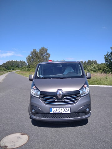 Szary bus Renault Trafic z czerwonym kogutem na dachu, zaparkowany na asfaltowej drodze w słoneczny dzień.