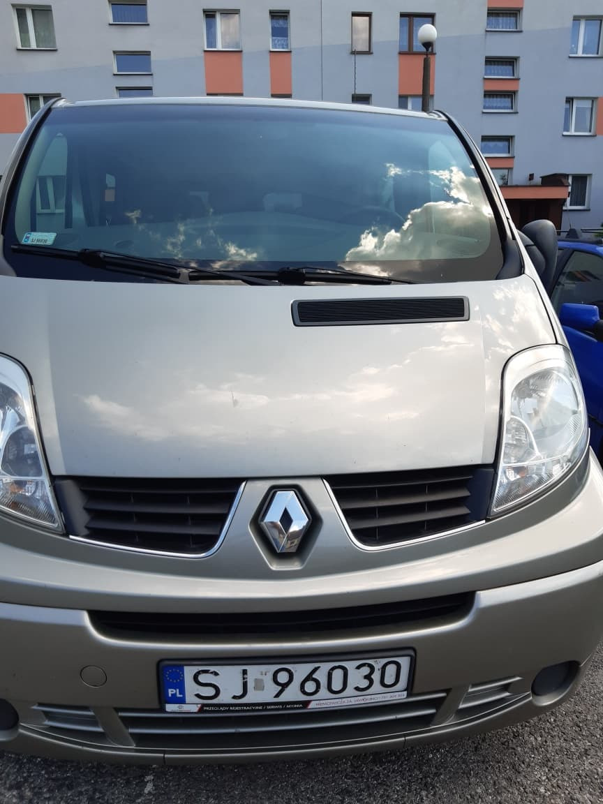 Przód srebrnego Renault Trafic z Jaworzna z widoczną tablicą rejestracyjną na tle bloku mieszkalnego. W szybie odbijają się chmury.