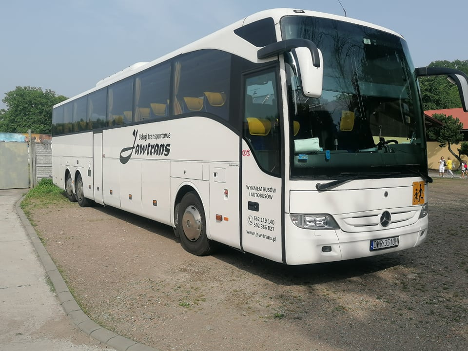 Biały autokar marki Mercedes z logo firmy Jawtrans, zaparkowany na żwirowym parkingu z widoczną naklejką informującą o wynajmie busów i autobusów oraz numerami telefonów.