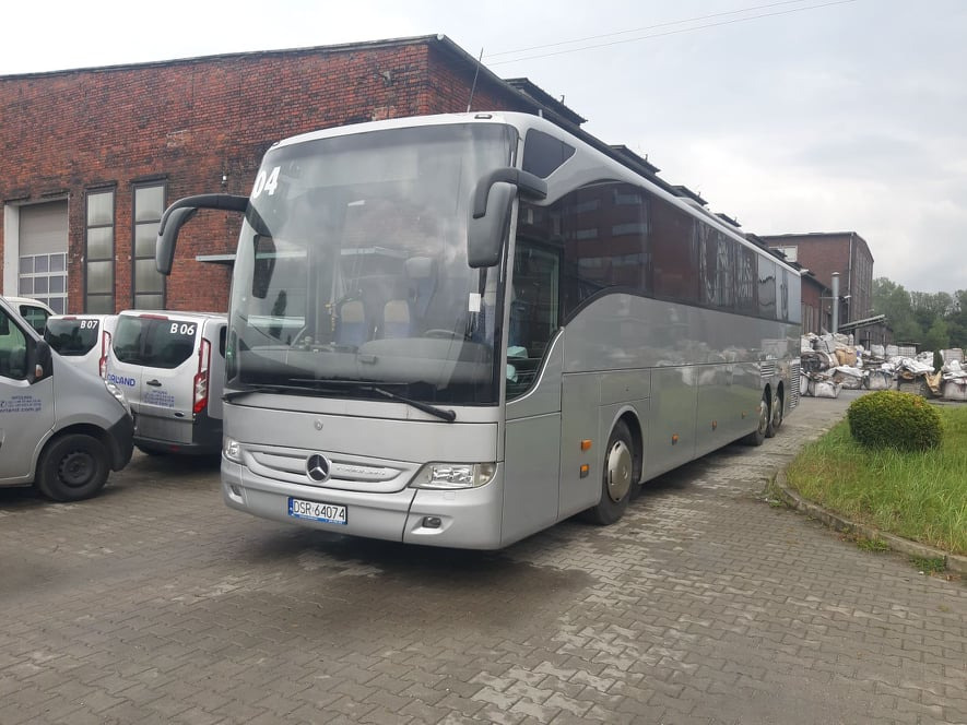 Srebrny autokar Mercedes-Benz z numerem bocznym '04' zaparkowany na brukowanym placu, z budynkiem z czerwonej cegły w tle i zaparkowanymi busami z logo firmy transportowej po lewej stronie.