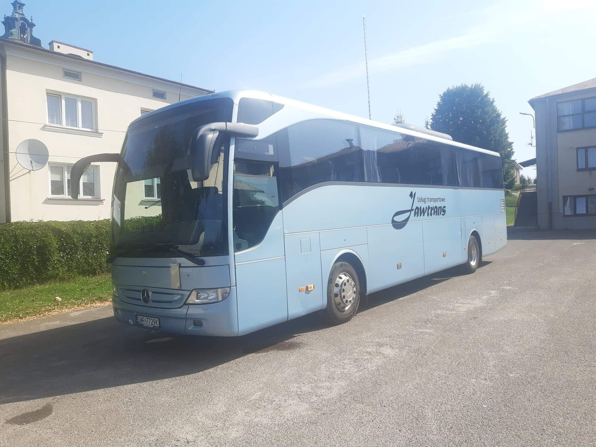 Błękitny autokar turystyczny marki Mercedes z logo firmy Awtrans na parkingu przed budynkiem w słoneczny dzień.