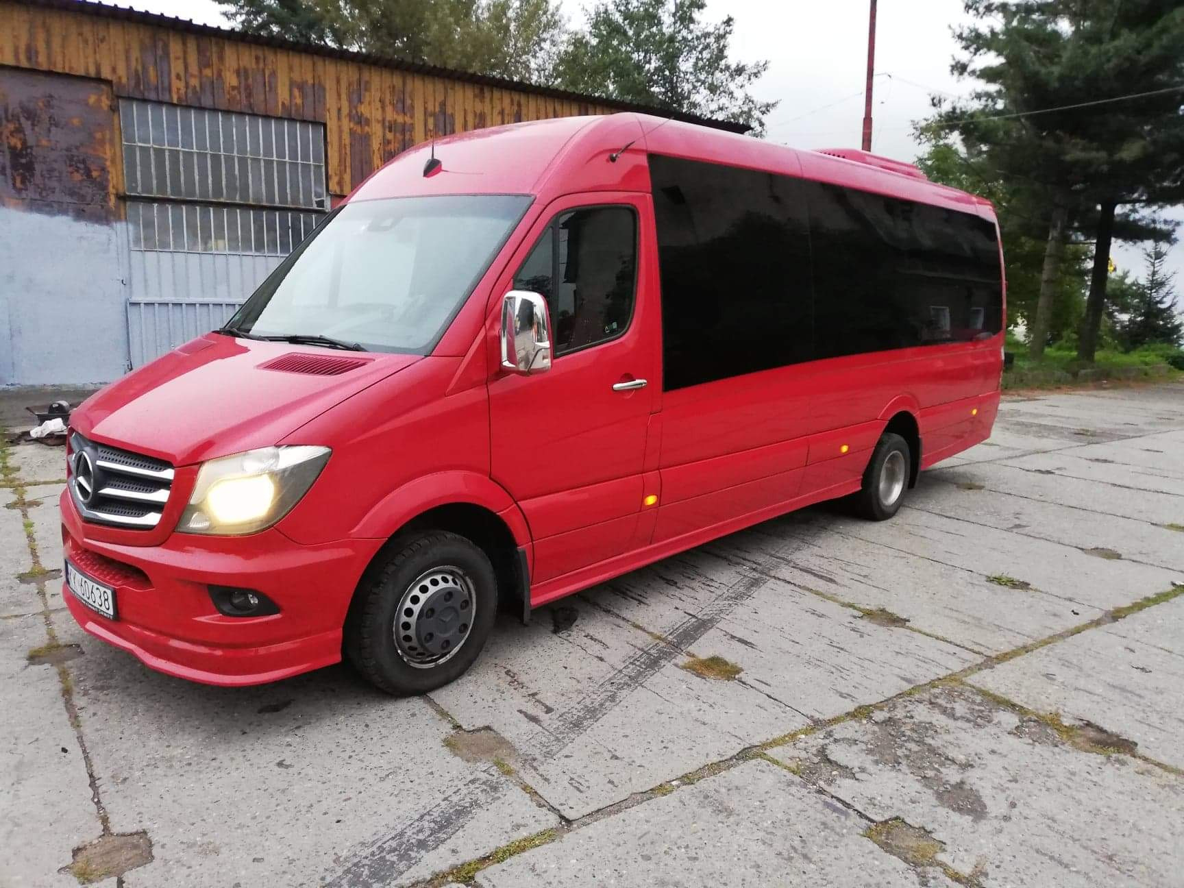 Czerwony bus Mercedes Sprinter z przyciemnianymi szybami, zaparkowany na betonowym placu przed budynkiem z blachy falistej.