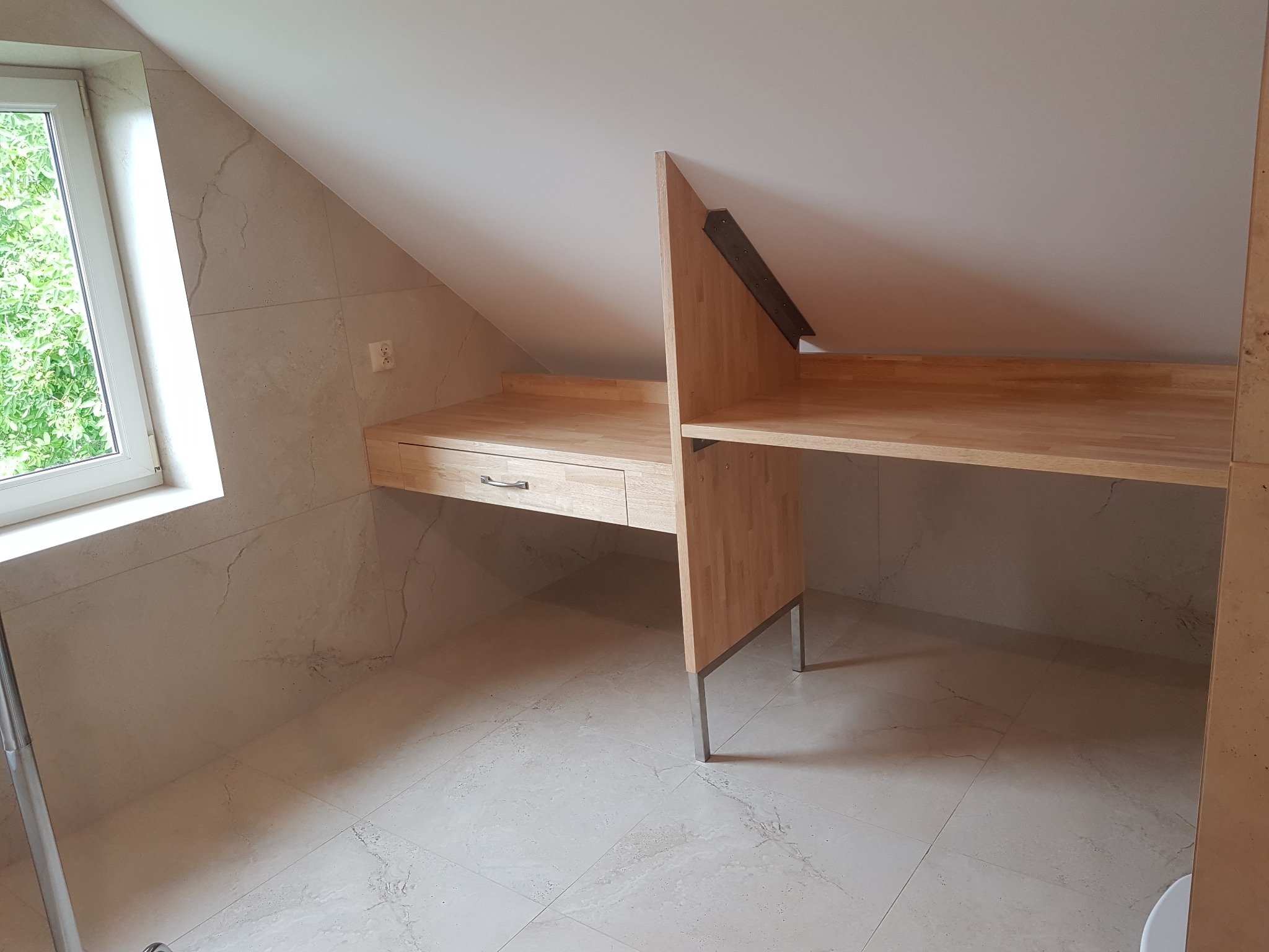 Drewniany blat biurka w jasnym odcieniu, zamontowany w pokoju na poddaszu, wsparty na metalowych nogach, z widocznymi płytkami imitującymi marmur na ścianach i podłodze.