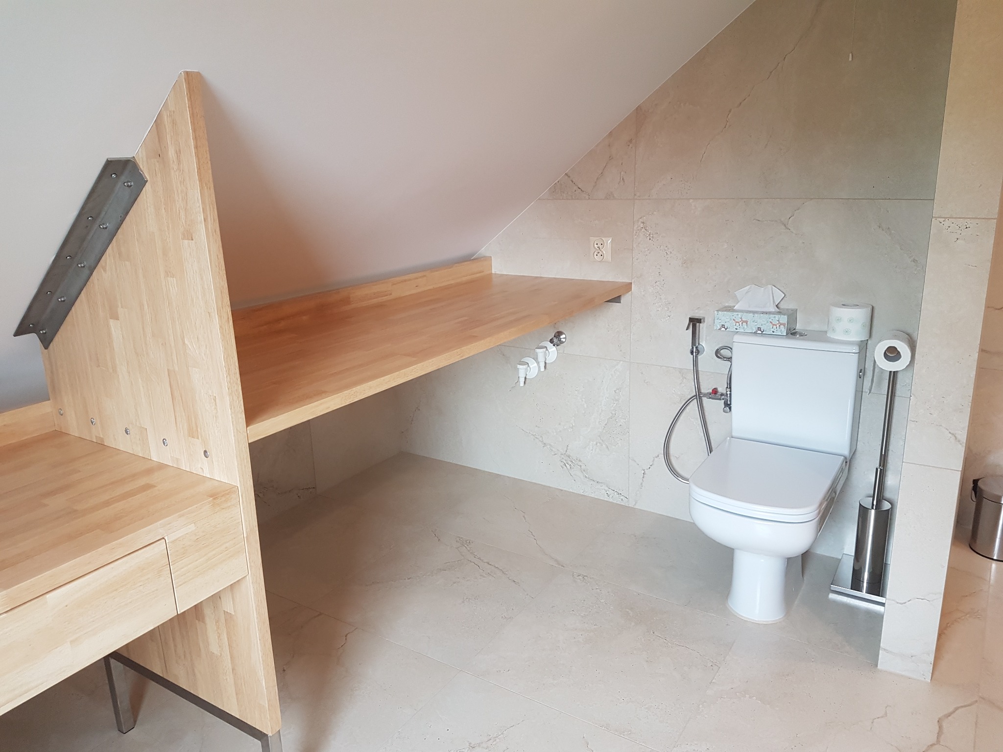 Jasna łazienka na poddaszu ze skośnym sufitem, z toaletą, drewnianym blatem roboczym zamontowanym na ścianie i drewnianą szafką z szufladą, wykończona płytkami imitującymi marmur.