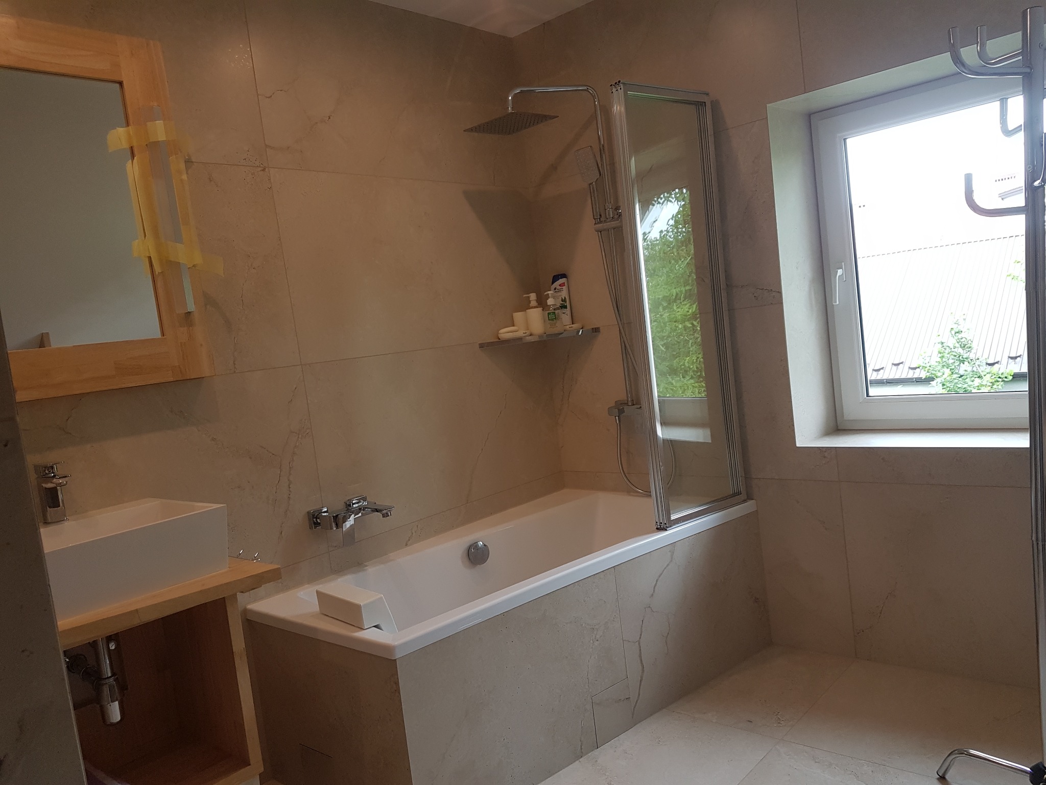 Wykończona łazienka w beżowych płytkach imitujących marmur, z wanną z parawanem nawannowym, umywalką nablatową z drewnianą szafką i lustrem zabezpieczonym taśmą malarską.