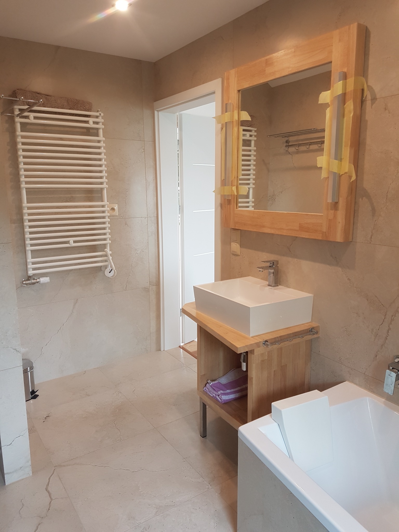 Wykończona łazienka w beżowych płytkach z drewnianą ramą lustra zabezpieczoną taśmą, białą umywalką na drewnianym blacie, grzejnikiem drabinkowym i fragmentem białej wanny.