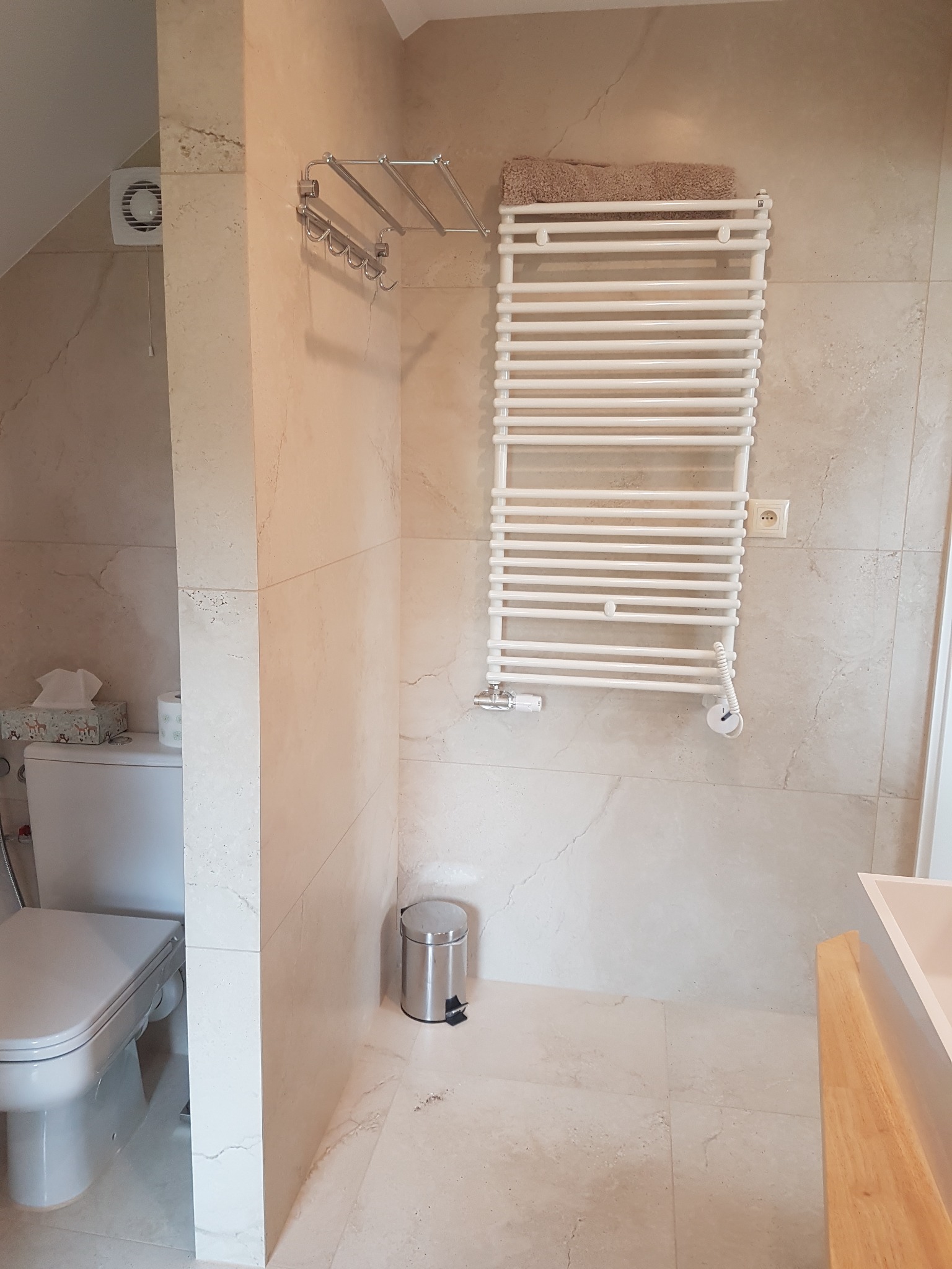 Jasna łazienka z beżowymi płytkami imitującymi marmur, widoczny grzejnik drabinkowy z ręcznikiem, toaleta z pudełkiem chusteczek i kosz na śmieci.