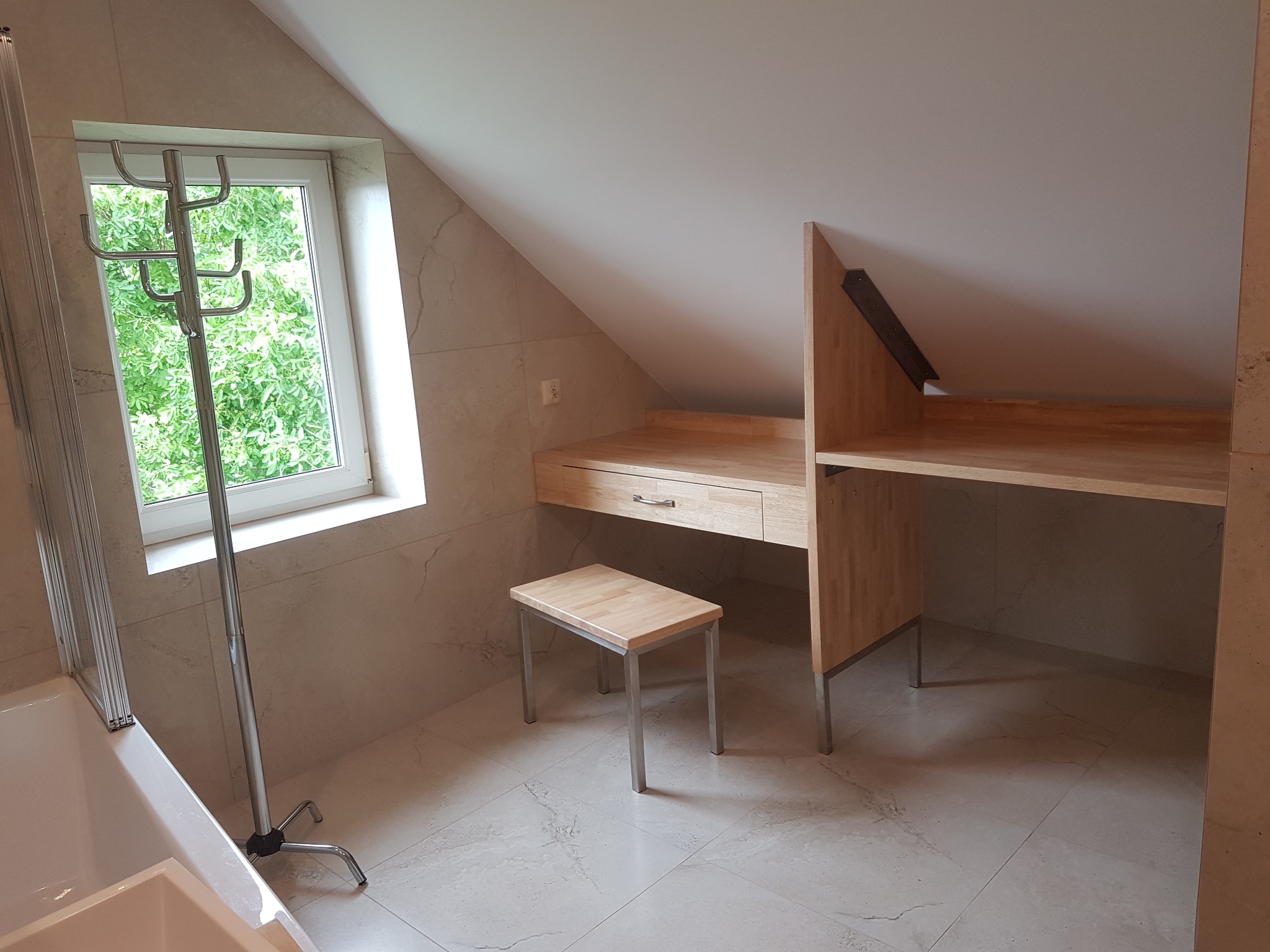 Jasna łazienka na poddaszu z beżowymi płytkami, drewnianym biurkiem z szufladą, stołkiem i chromowanym wieszakiem na ręczniki przy oknie.