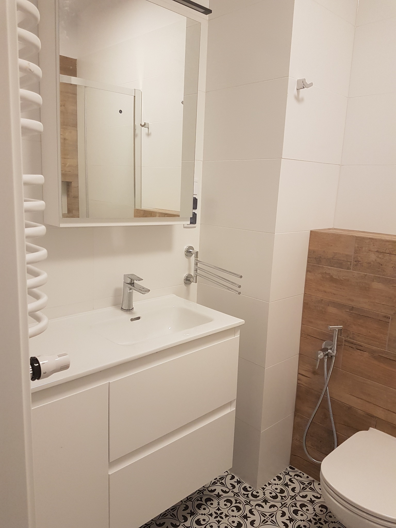 Nowoczesna łazienka po remoncie: białe płytki, umywalka z szafką, lustro, dekoracyjne płytki podłogowe w stylu retro, drewnopodobna okładzina ściany z bidetką.