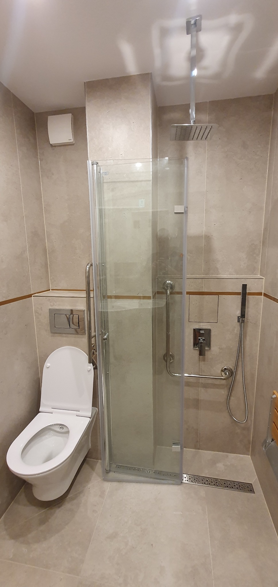 Wykończona łazienka z beżowymi płytkami, kabiną prysznicową z uchwytami dla niepełnosprawnych, podwieszaną toaletą i odpływem liniowym.