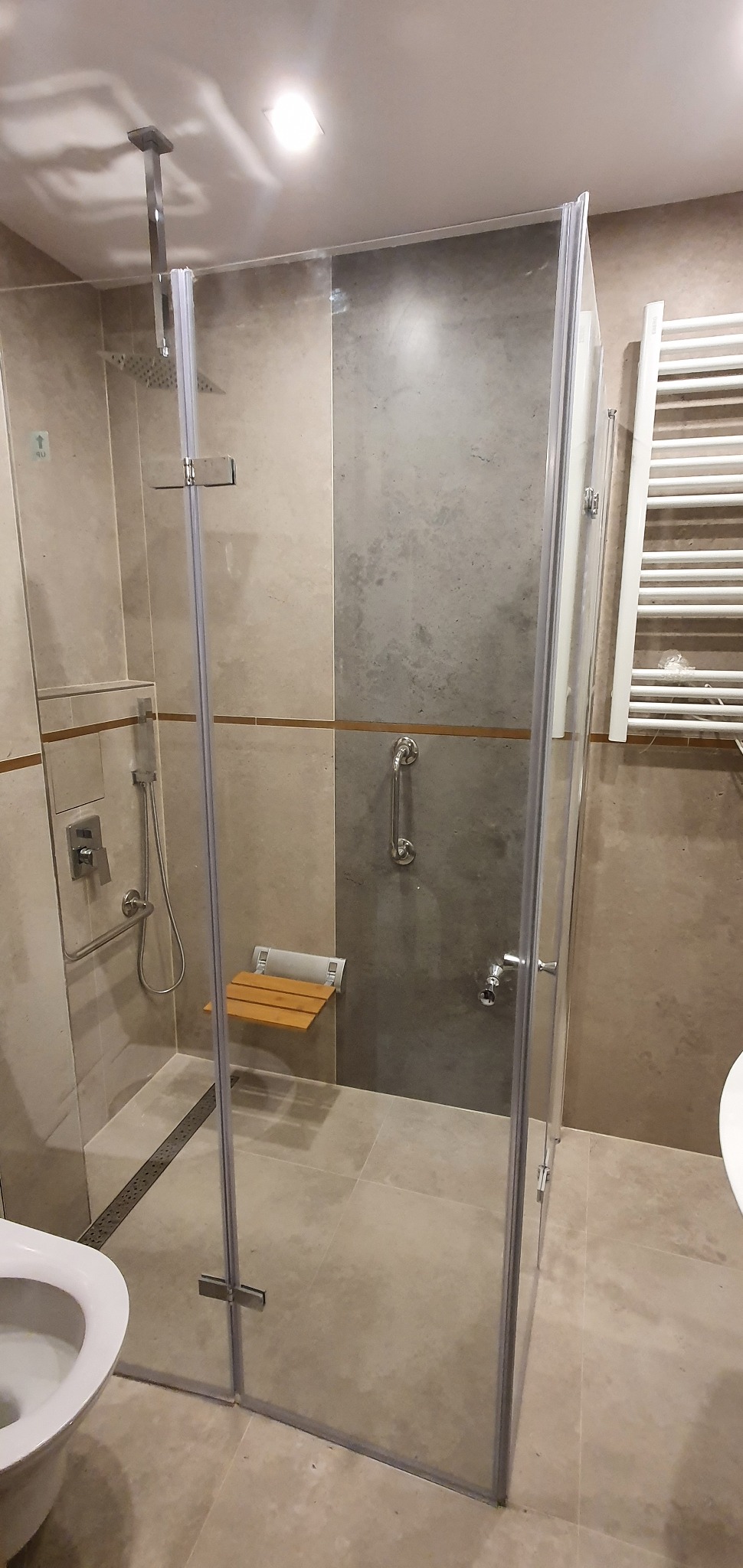 Wykończona łazienka z kabiną prysznicową ze szklanymi ściankami, zamontowanym siedziskiem pod prysznicem i uchwytami dla osób z niepełnosprawnościami, widoczny również grzejnik drabinkowy.