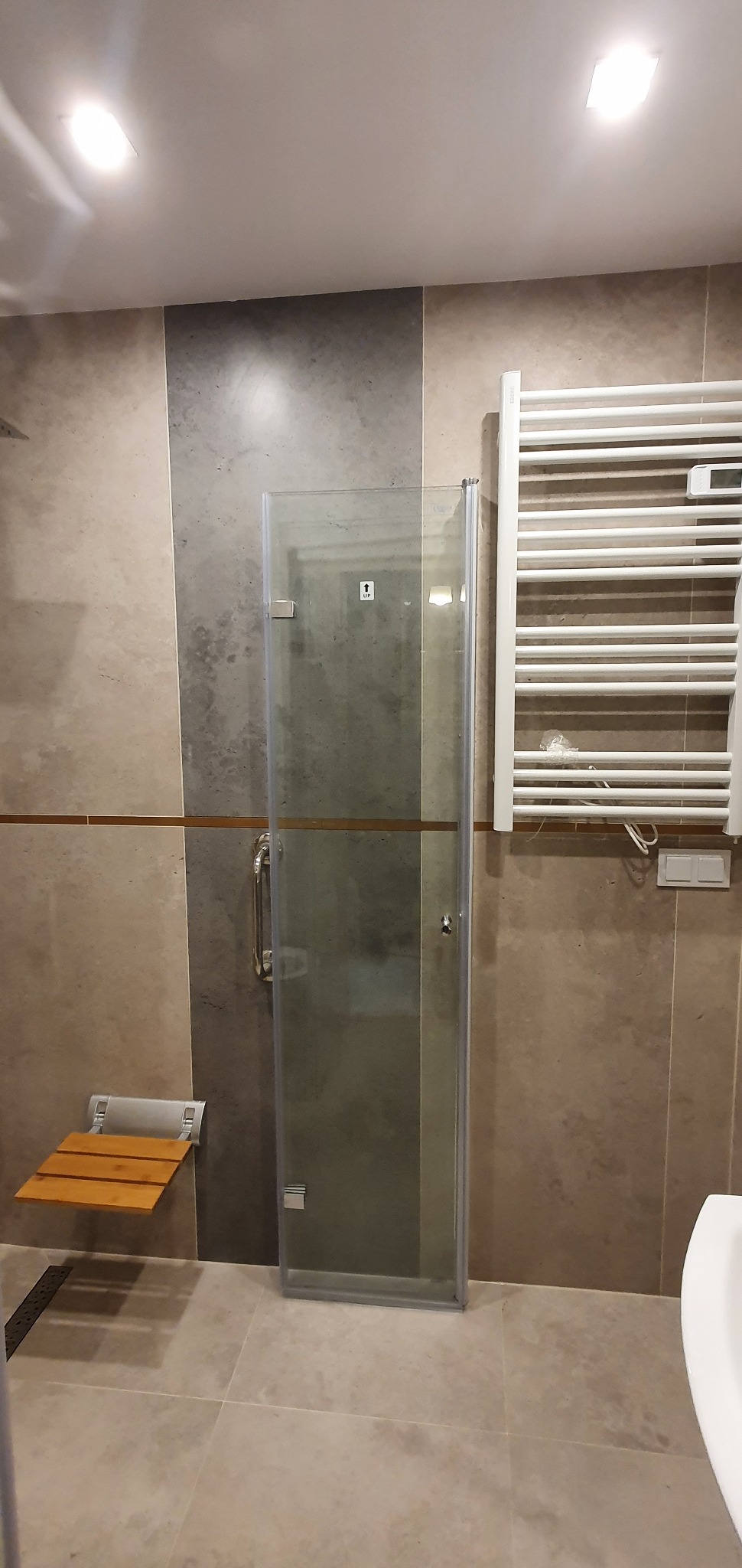 Zamknięta szklana kabina prysznicowa z metalowym uchwytem i naklejką informacyjną, zamontowana w łazience wykończonej płytkami w odcieniach beżu i szarości, z drewnianym, składanym siedziskiem...