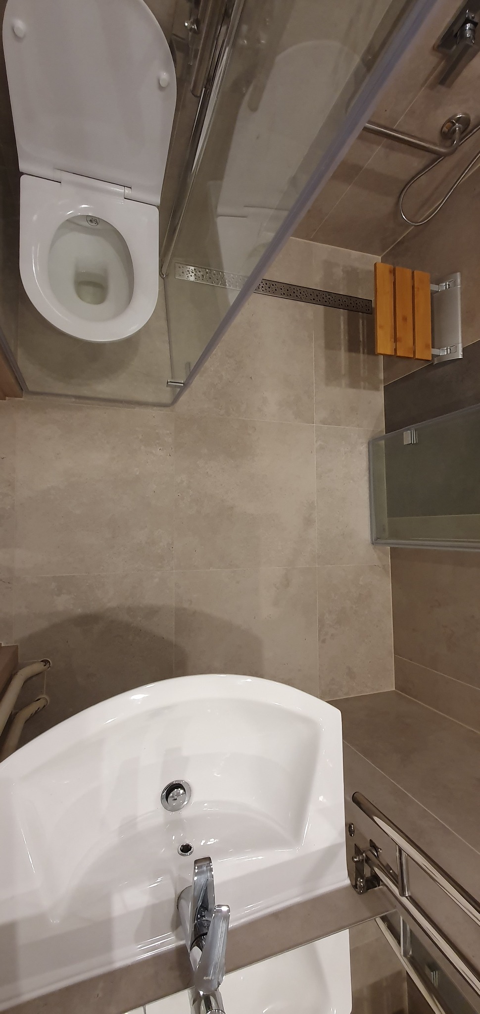 Widok z góry na wykończoną łazienkę z beżowymi płytkami, białą umywalką i toaletą wiszącą, szklaną kabiną prysznicową z drewnianym siedziskiem.