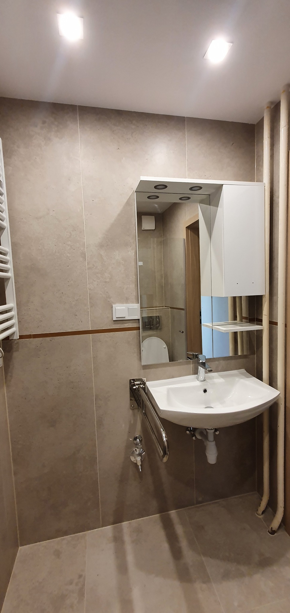 Umywalka narożna zamontowana na chromowanym wsporniku z uchwytem dla niepełnosprawnych, nad nią lustro z szafką i oświetleniem punktowym, beżowe płytki na ścianach i podłodze, widoczne rury grzewcze.