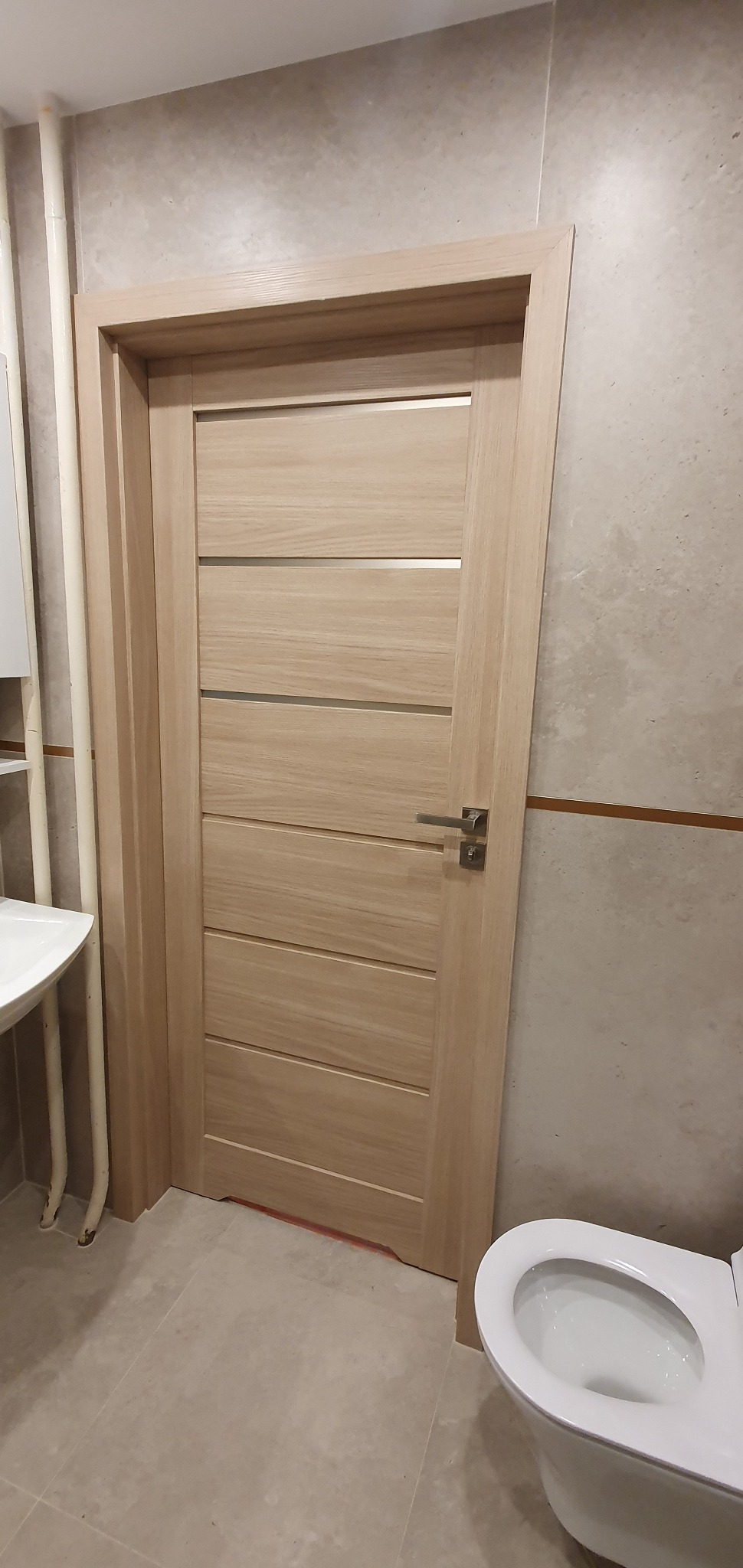 Jasna, drewniana, wewnętrzna ościeżnica z zamontowanymi drzwiami w łazience z beżowymi płytkami, widoczna umywalka i toaleta.