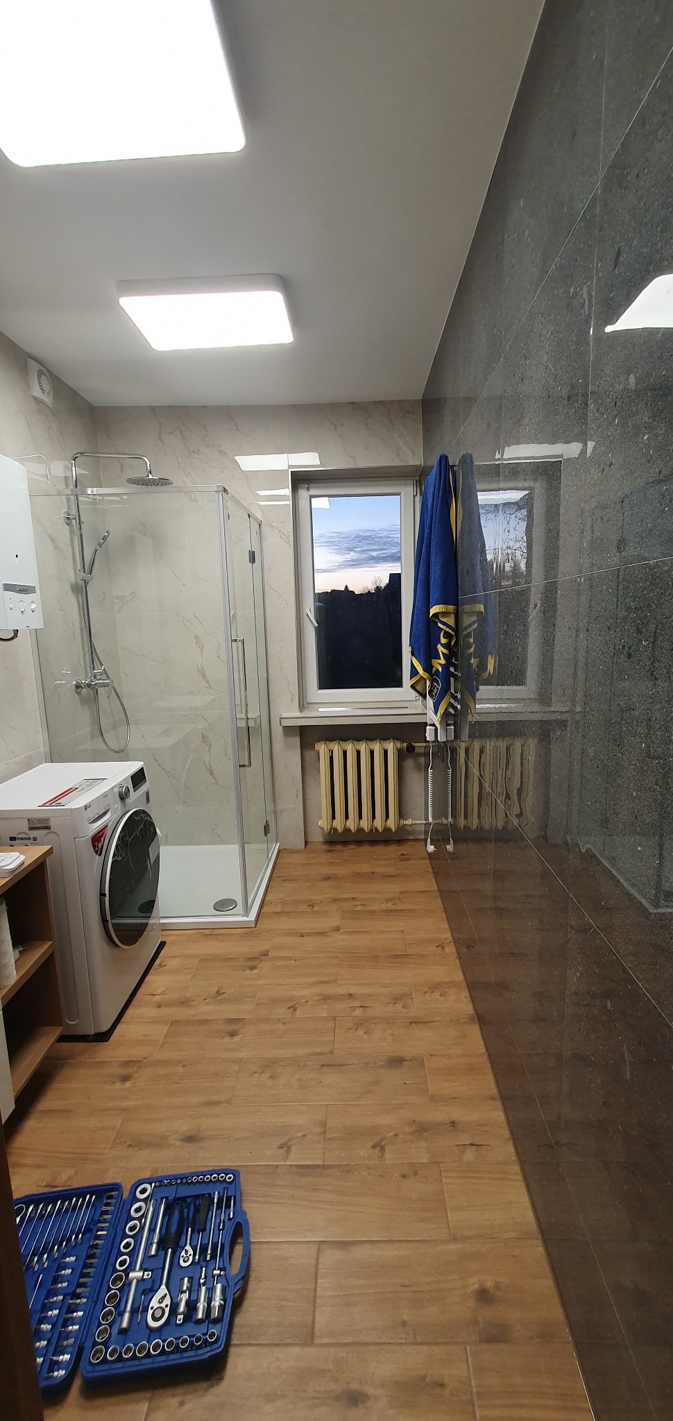 Wykończona łazienka z kabiną prysznicową, pralką i grzejnikiem, na podłodze panele imitujące drewno, a na ścianie szare płytki, w oknie widok na zachodzące słońce, na podłodze leży zestaw narzędzi.