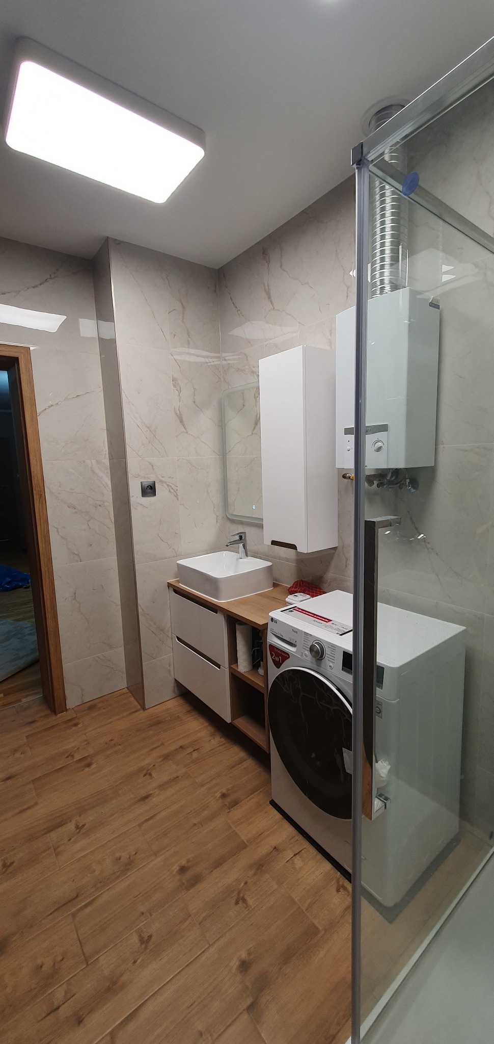 Nowoczesna łazienka z marmurowymi płytkami, drewnianą podłogą, umywalką nablatową, szafką wiszącą, pralką i kabiną prysznicową ze szklaną ścianką.