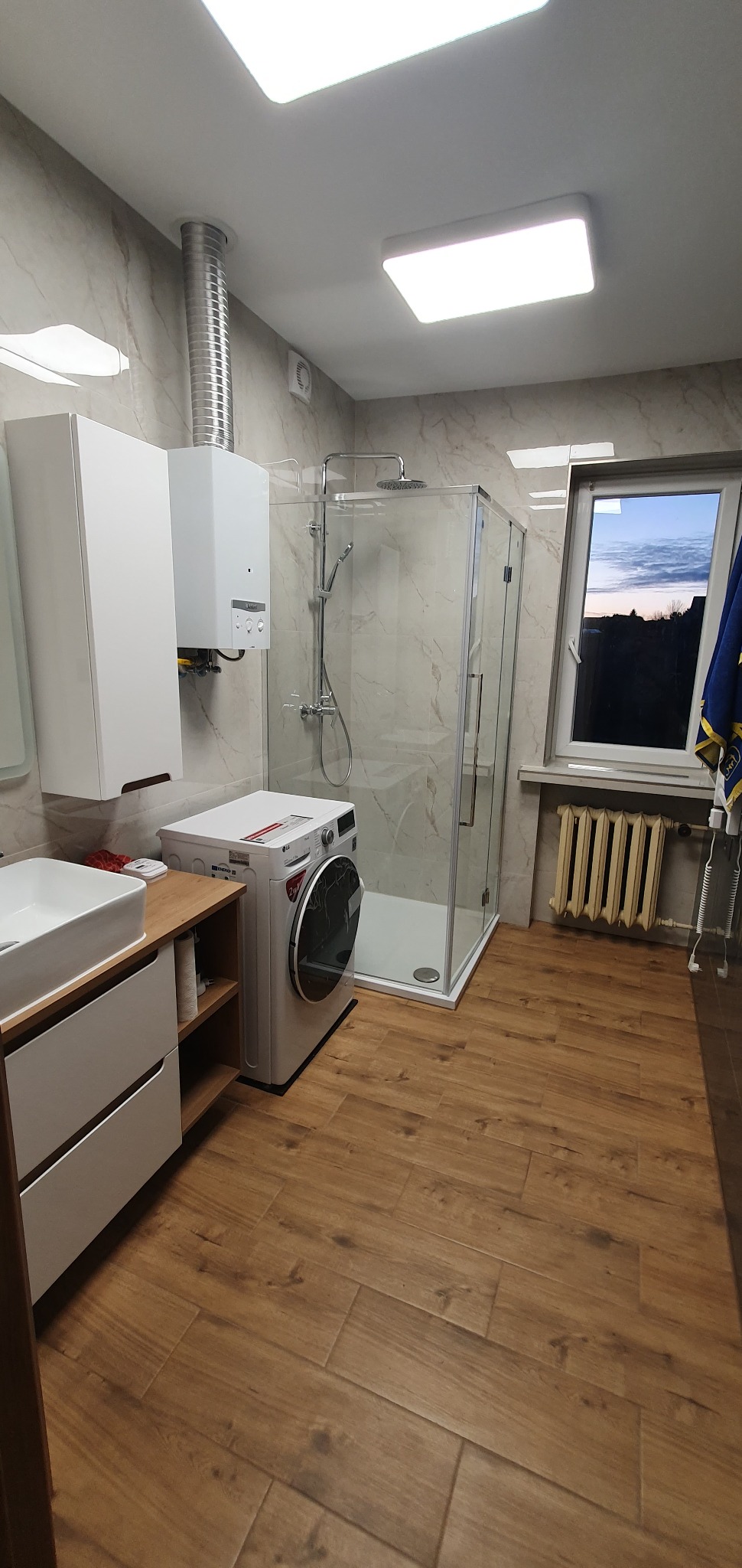 Nowoczesna łazienka z kabiną prysznicową, pralką LG, umywalką nablatową na drewnianym blacie, grzejnikiem żeberkowym pod oknem i marmurowymi płytkami na ścianach.