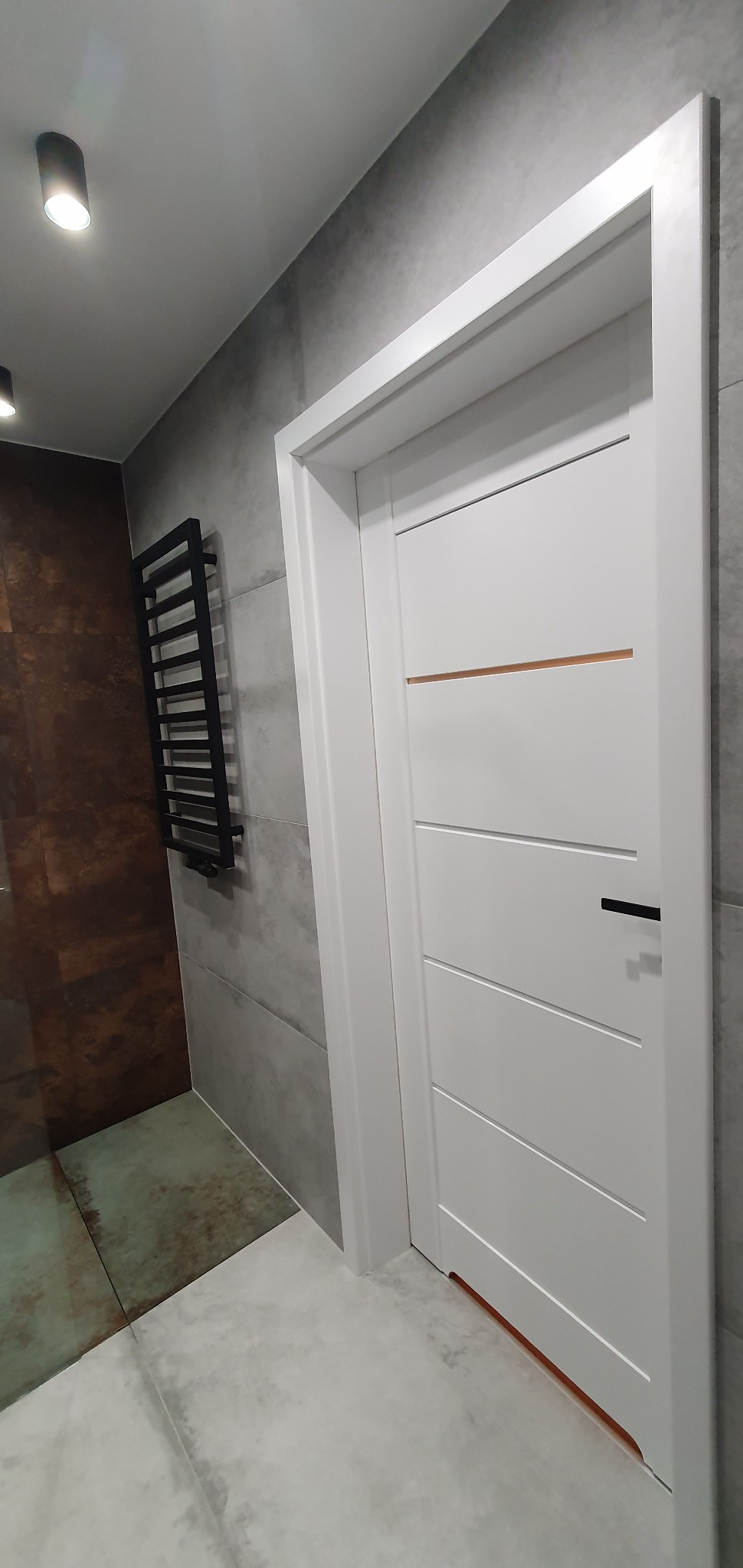 Nowoczesna łazienka z białą, minimalistyczną stolarką drzwiową z poziomymi frezowaniami, czarnym uchwytem i subtelnym, miedzianym akcentem, na tle szarych płytek imitujących beton i czarnego...