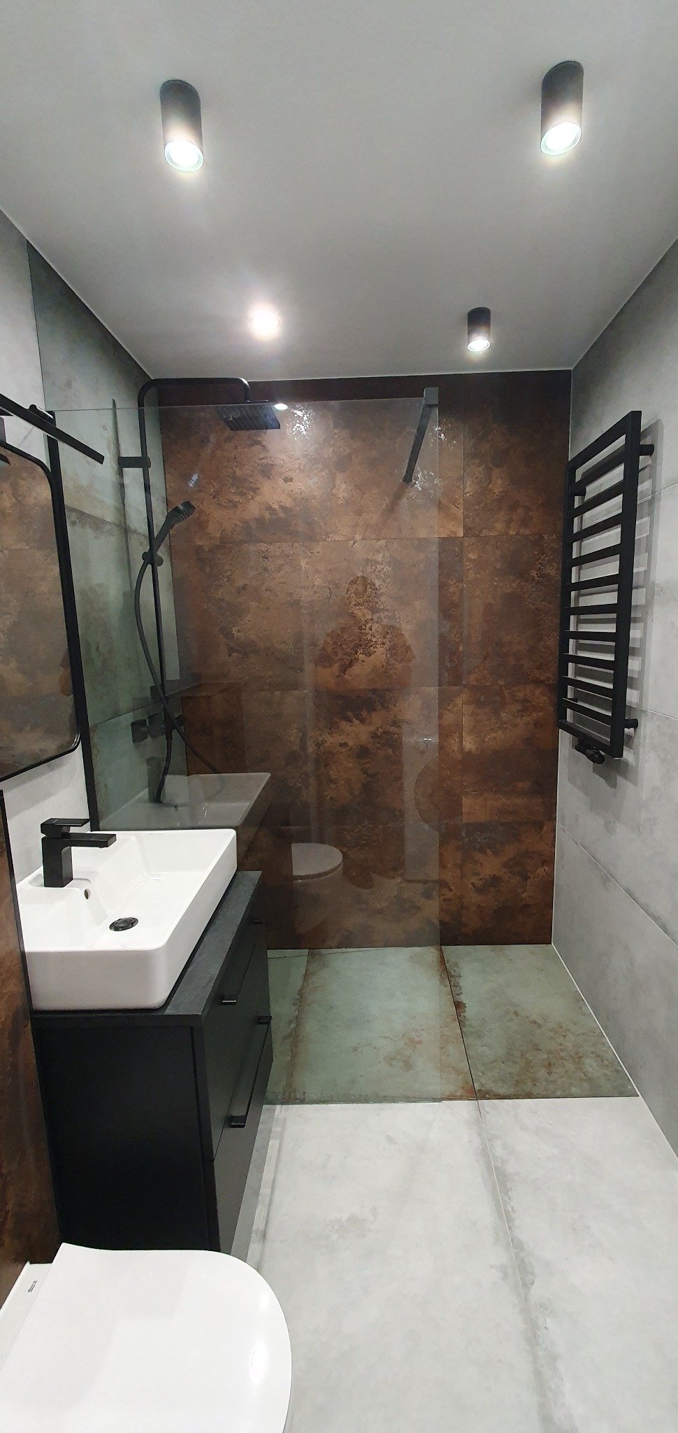 Nowoczesna łazienka z kabiną prysznicową typu walk-in, umywalką nablatową na czarnej szafce, grzejnikiem drabinkowym w kolorze czarnym oraz płytkami imitującymi rdzę na ścianie prysznica.