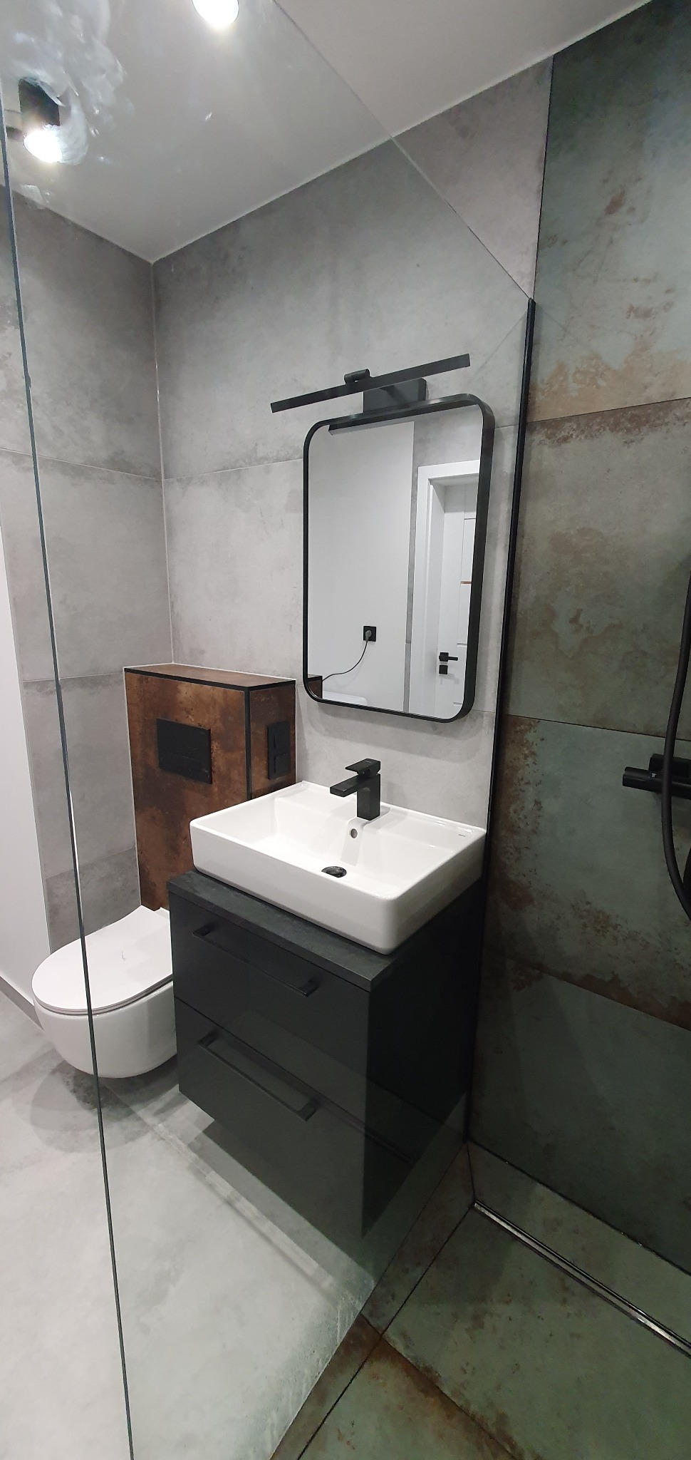 Nowoczesna łazienka z umywalką nablatową na czarnej szafce z szufladami, toaletą w zabudowie z obudową imitującą rdzę oraz lustrem z czarną ramą i oświetleniem LED.