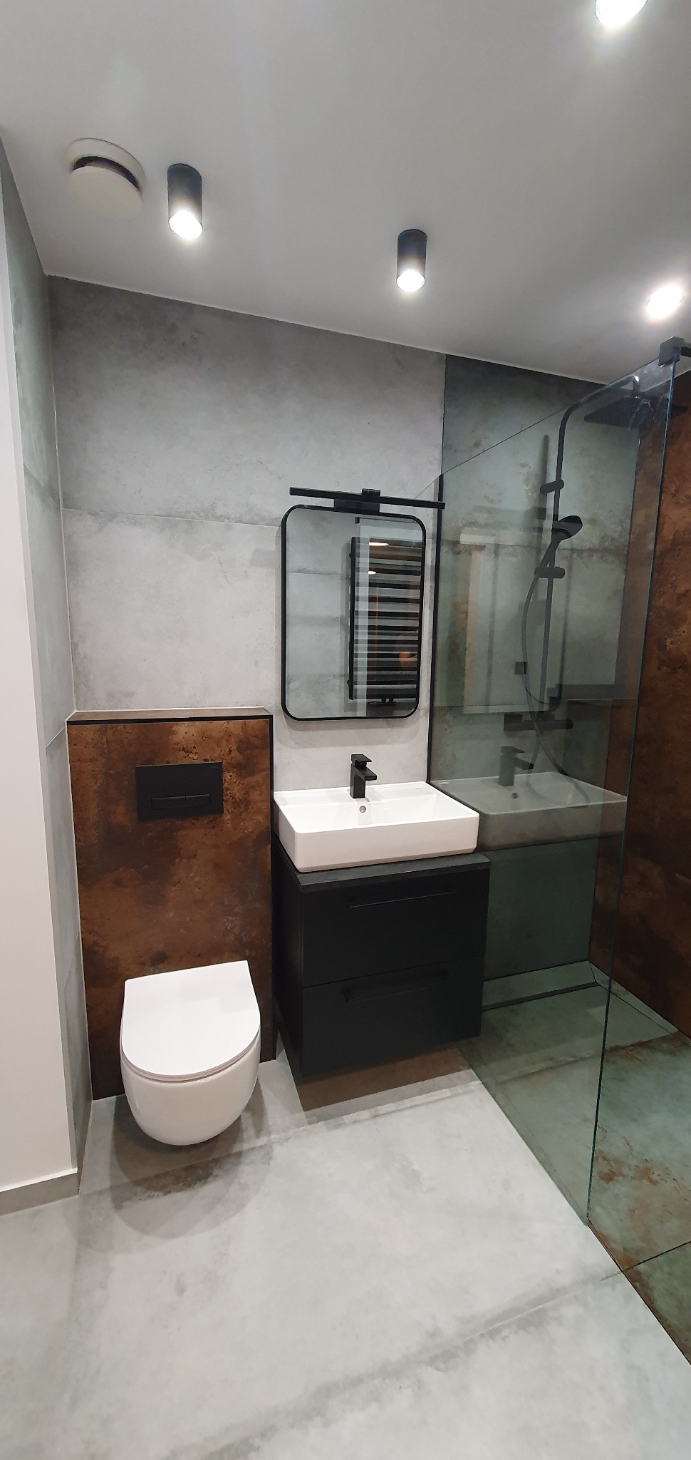 Nowoczesna łazienka z betonowymi płytkami, wiszącą toaletą, czarną szafką podumywalkową z kwadratową umywalką i kabiną prysznicową ze szklaną ścianką.