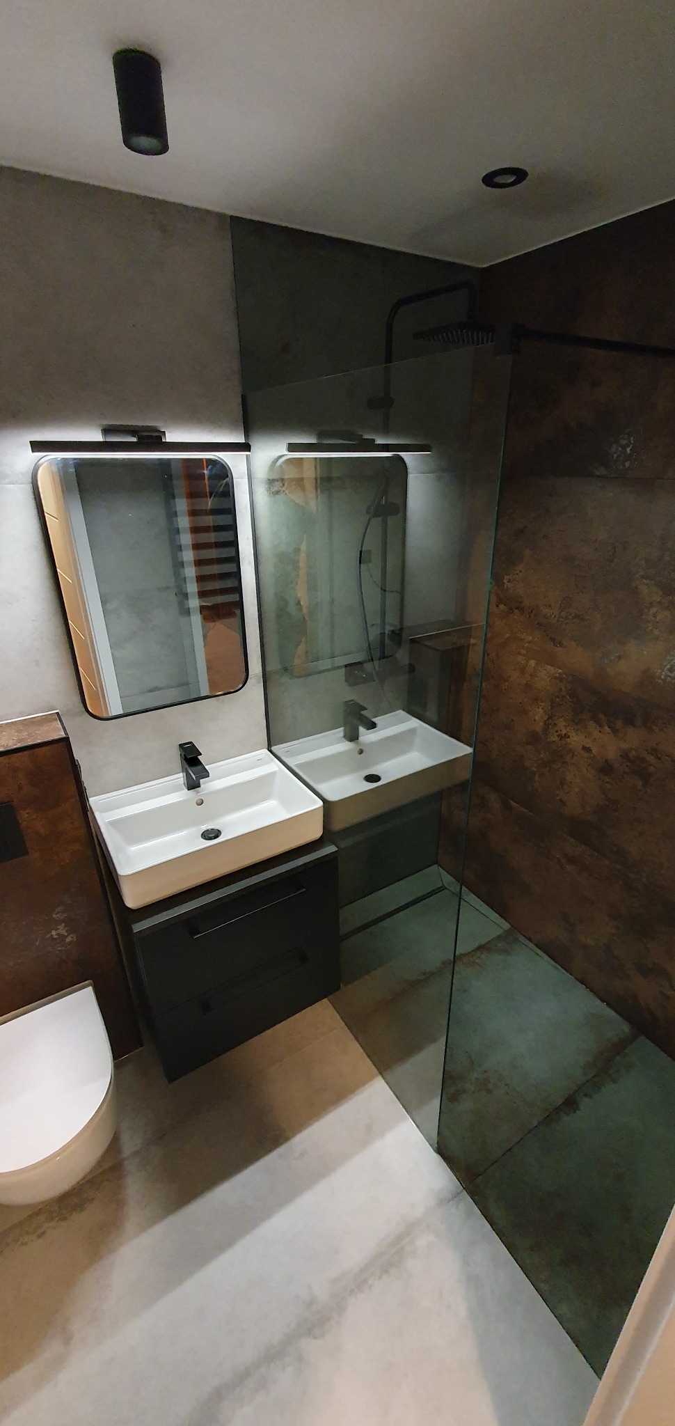 Nowoczesna łazienka z dwiema umywalkami nablatowymi na czarnych szafkach, lustrami z oświetleniem LED, kabiną prysznicową typu walk-in i płytkami imitującymi beton.