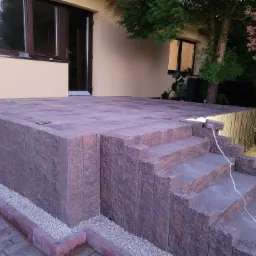 Wykończony taras i schody z kostki brukowej w odcieniach brązu, z widocznymi detalami tekstury kamienia, obok domu z beżową elewacją i oknami w ciemnych ramach, z dodatkowym oświetleniem...