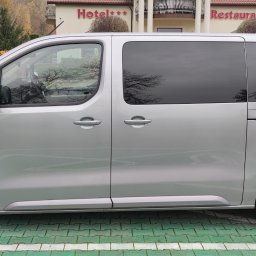 Georgetravel Katarzyna Czerewko-Pandya Grzegorz Ozimek Spółka Cywilna - Srebrny, elektryczny van zaparkowany na stacji ładowania przed budynkiem z napisem Hotel i Restauracja, widok z boku.