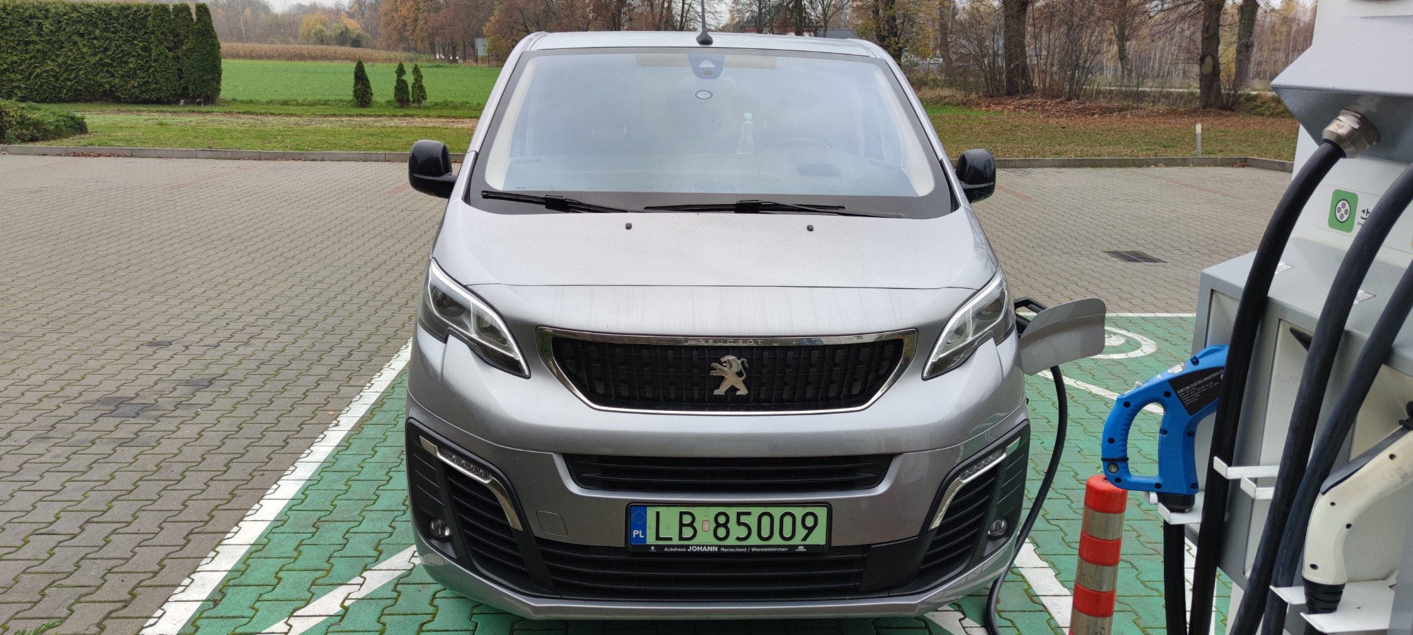 Srebrny, elektryczny Peugeot Expert zaparkowany na stanowisku ładowania z zielonymi oznaczeniami, podłączony do stacji ładowania, na tle zielonego trawnika i drzew z jesiennymi liśćmi.