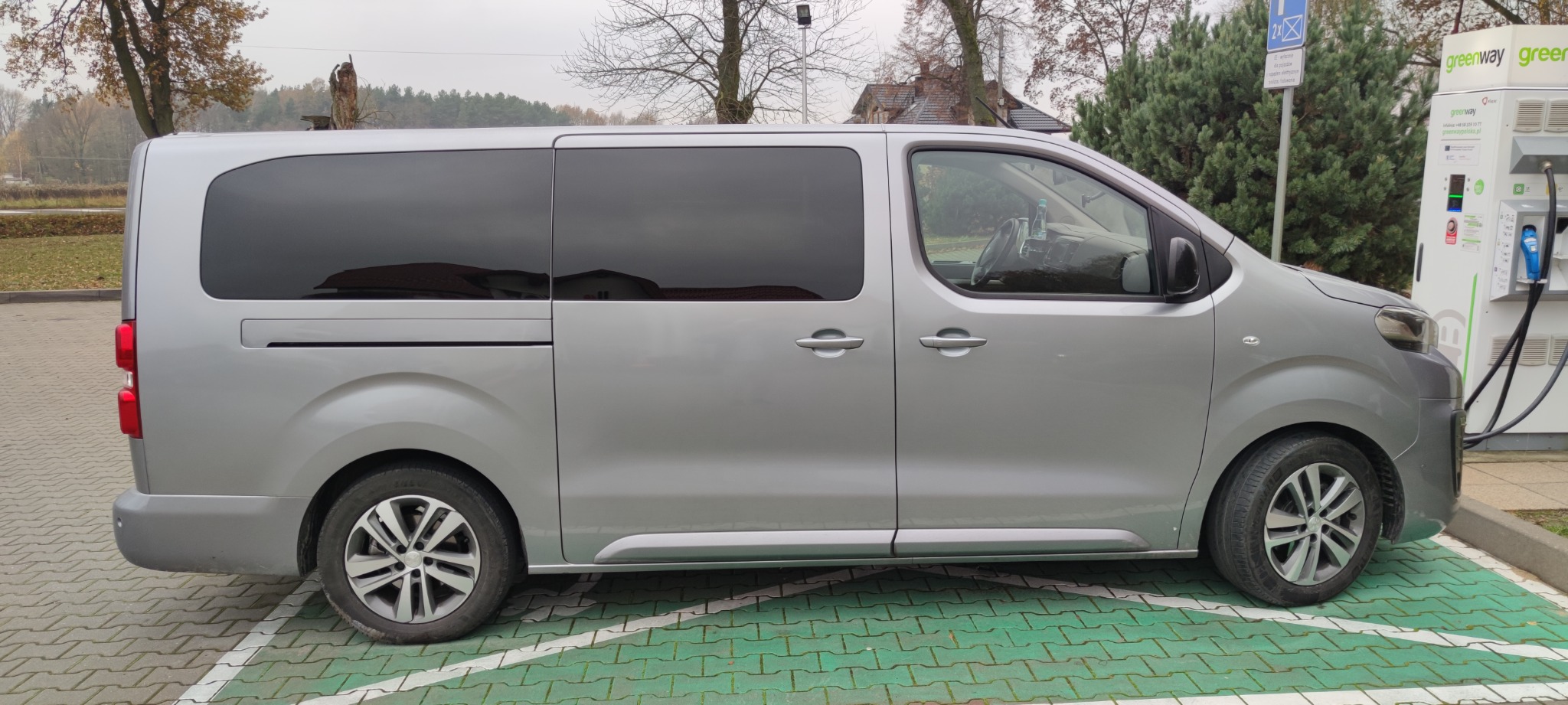 Srebrny van z przyciemnianymi szybami zaparkowany przy stacji ładowania pojazdów elektrycznych Greenway, widok z boku na tle drzew bez liści.