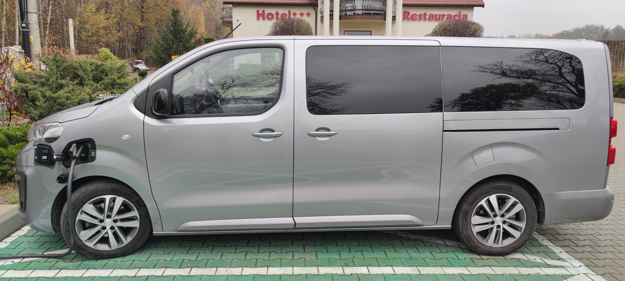 Srebrny, elektryczny van zaparkowany na stacji ładowania przed budynkiem z napisem Hotel i Restauracja, widok z boku.