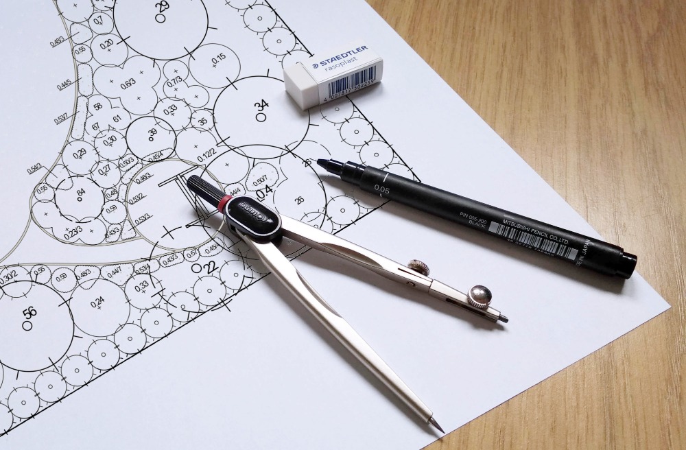 Rzut ogrodu z precyzyjnie oznaczonymi obszarami nasadzeń, leżący na drewnianym blacie obok cyrkla, cienkopisu i gumki do mazania Staedtler.