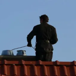 Osoba w czarnym kombinezonie na dachu, z narzędziem do czyszczenia komina, widok z tyłu, na tle błękitnego nieba.