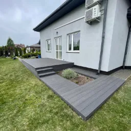 Szary taras kompozytowy z trzema schodkami, otaczający rabatę z rośliną, przylegający do jasnego budynku z dwoma klimatyzatorami na ścianie.