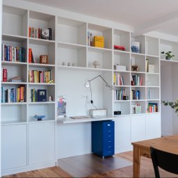Jurek Popowicz - Białe regały na książki z wbudowanym biurkiem i niebieską szafką z szufladami. Na stole wazon z kwiatami. Jasne wnętrze z drewnianą podłogą.