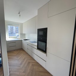 Jurek Popowicz - Minimalistyczna kuchnia z frontami w odcieniach bieli, czarnym sprzętem AGD i parkietem w jodełkę. Widok na okno z widokiem na budynek w rusztowaniach.