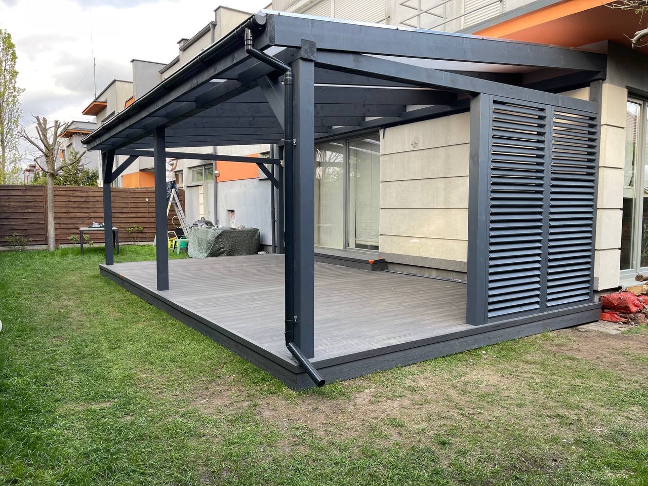 Zadaszony taras z szarej deski kompozytowej, z elementami drewnianymi pomalowanymi na ciemny szary kolor, przylegający do nowoczesnego domu z szarą elewacją i oknami przesuwnymi.