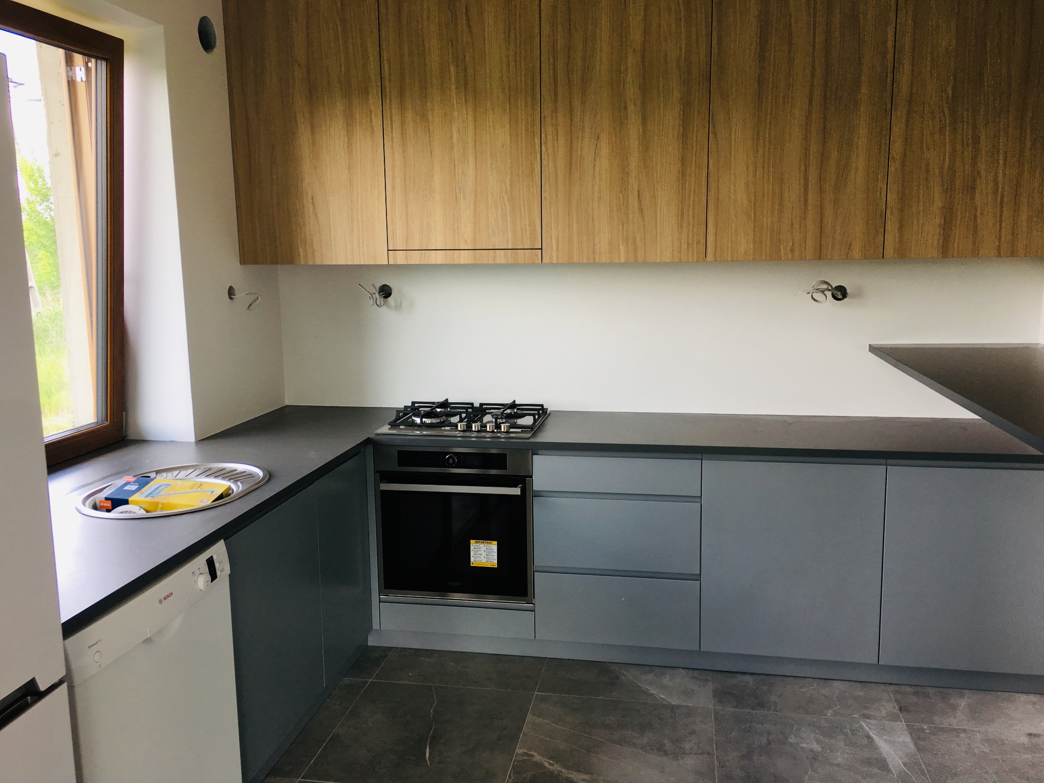 Nowoczesna kuchnia z szarymi szafkami dolnymi i górnymi szafkami z drewna, płyta gazowa i piekarnik w zabudowie, zlew z ociekaczem i akcesoriami, szare płytki na podłodze, widok z przodu.