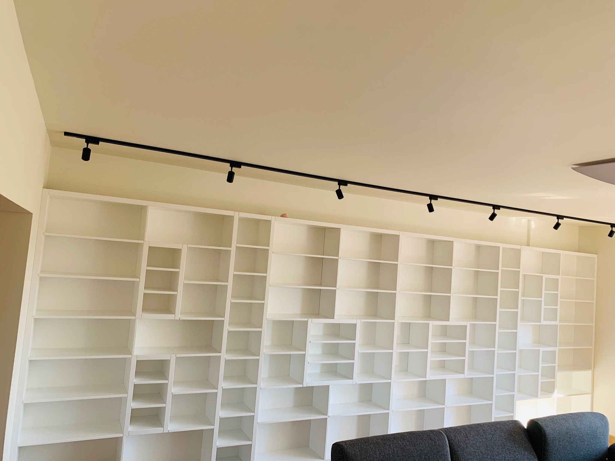Biała, modułowa biblioteka na całą ścianę z oświetleniem punktowym na szynie, widok z dołu, fragment szarej sofy na pierwszym planie.